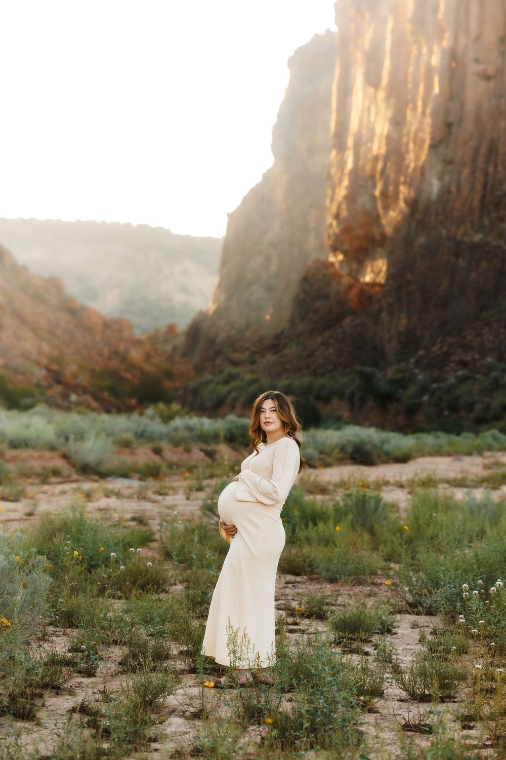 Pregnant woman in cream dress standing in desert canyon landscape at golden hour.