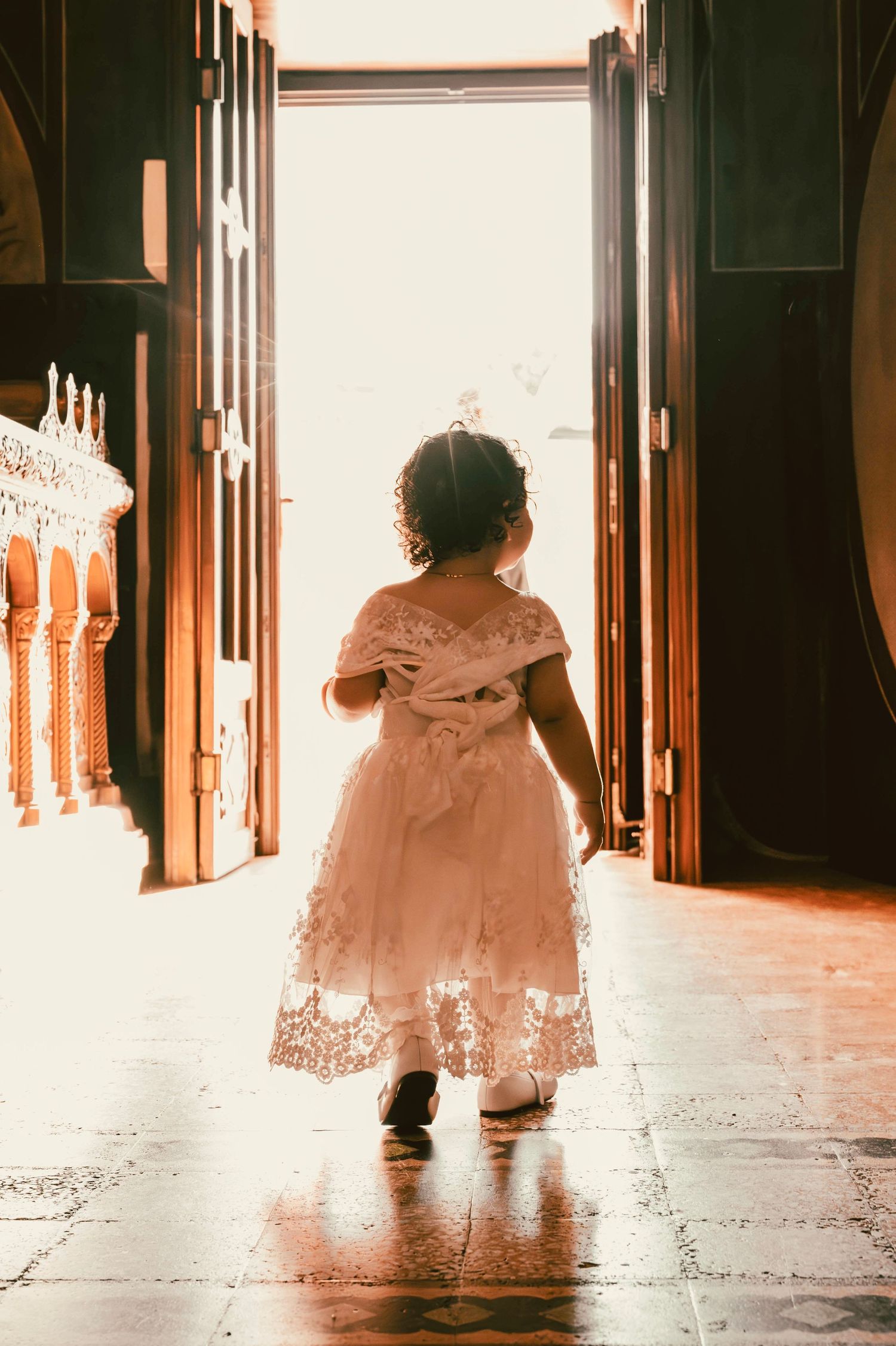 A small child in a lacy dress walking through ornate wooden doors into bright light.