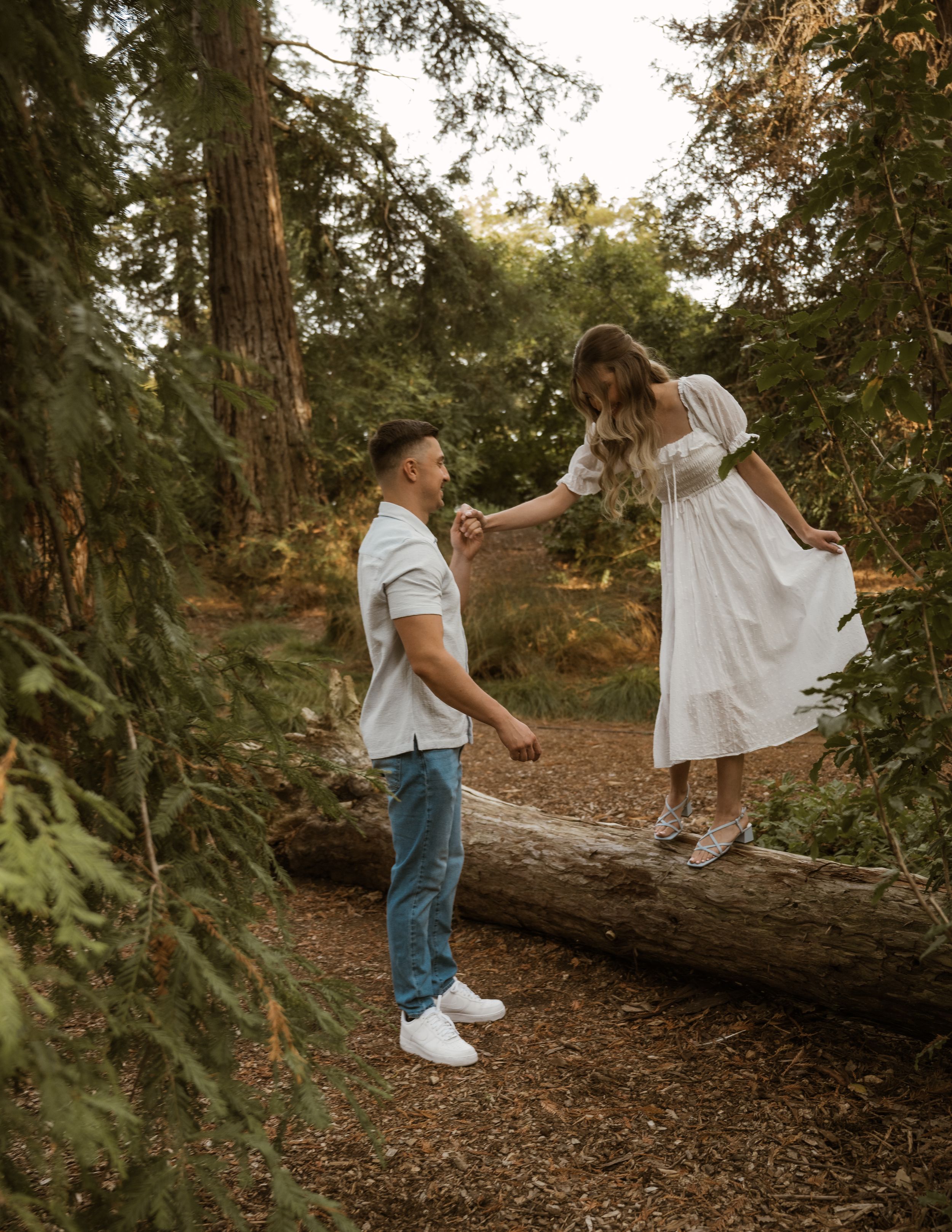 A couple plays and balances on a fallen log in a forest setting.