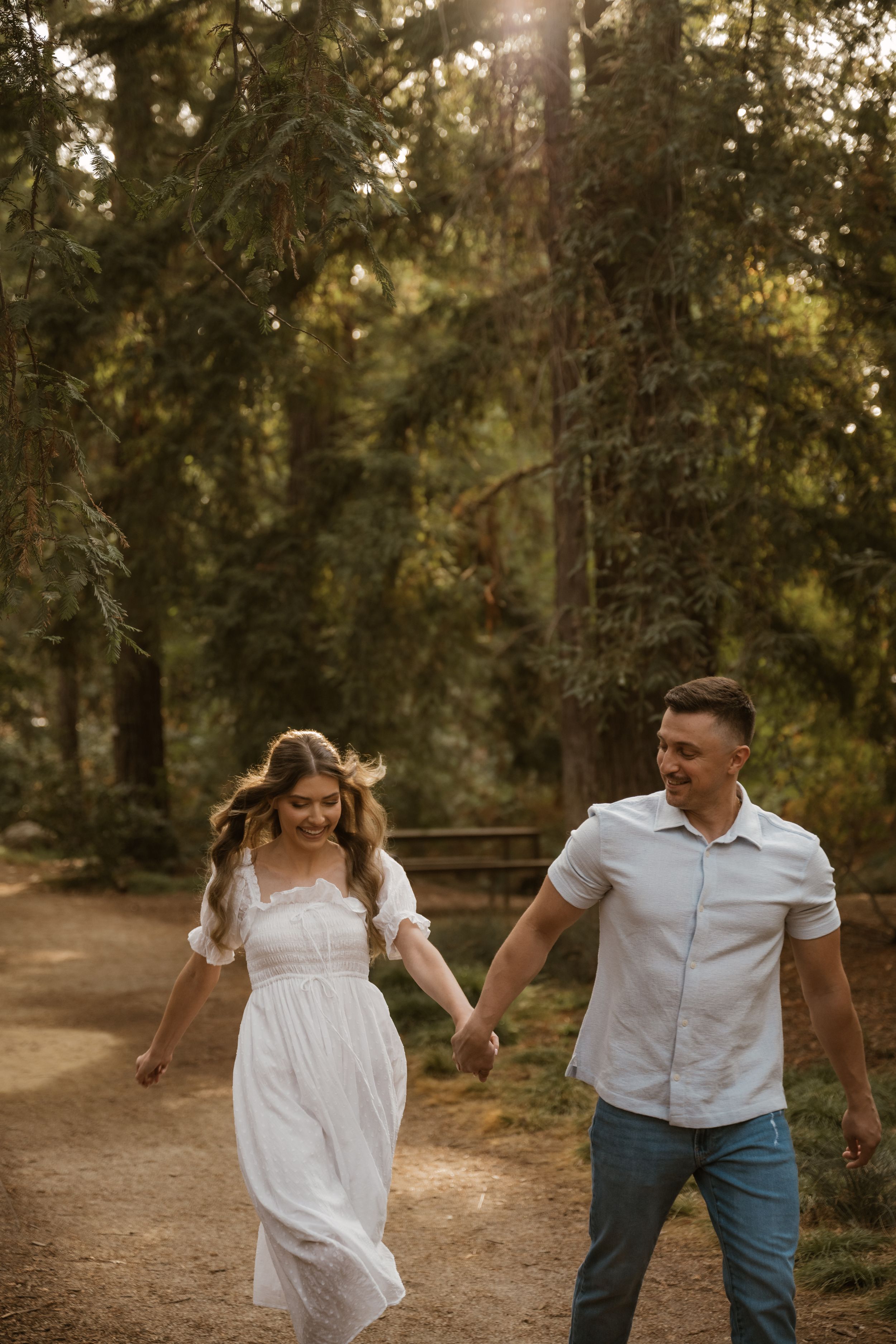 A couple in white and blue outfits playfully dance together in a sunlit forest setting.