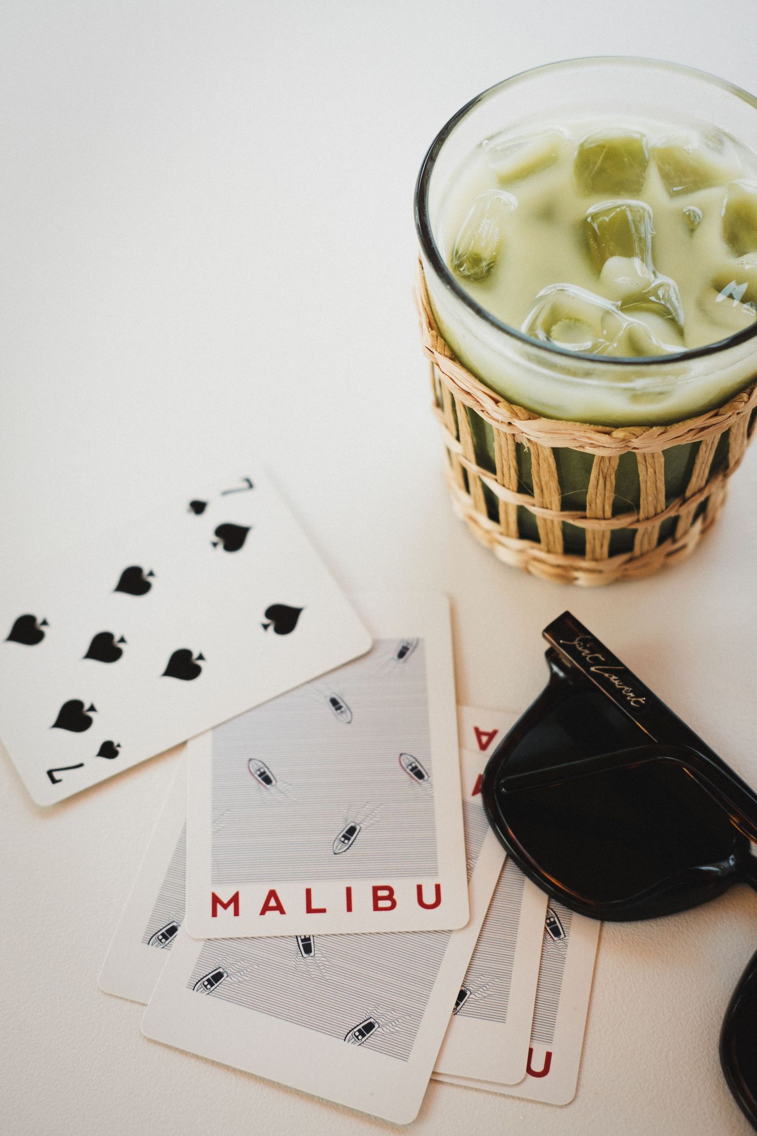 Playing cards and a Malibu bottle on a white surface with a woven glass holder.