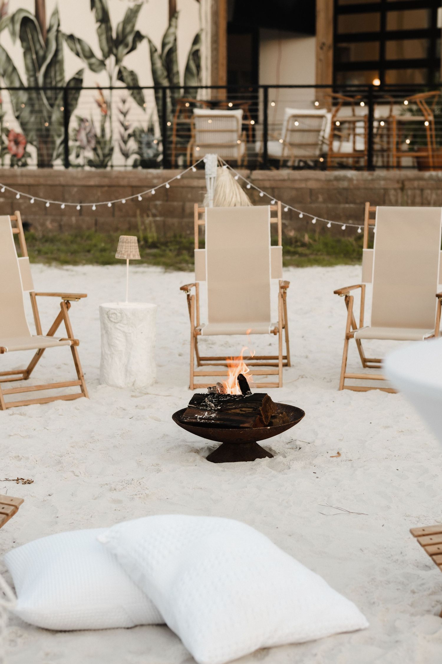 Beach lounge setup with white pillows and wooden chairs on sand under string lights.