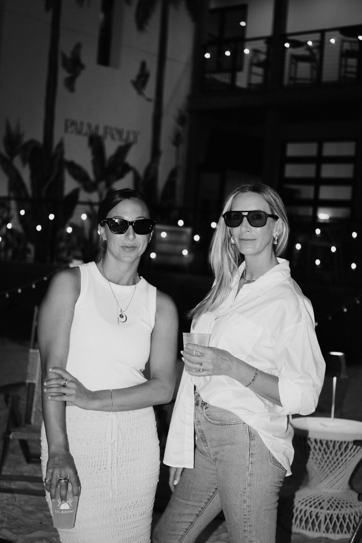 Two women in white tops and sunglasses pose together in a casual summer setting.