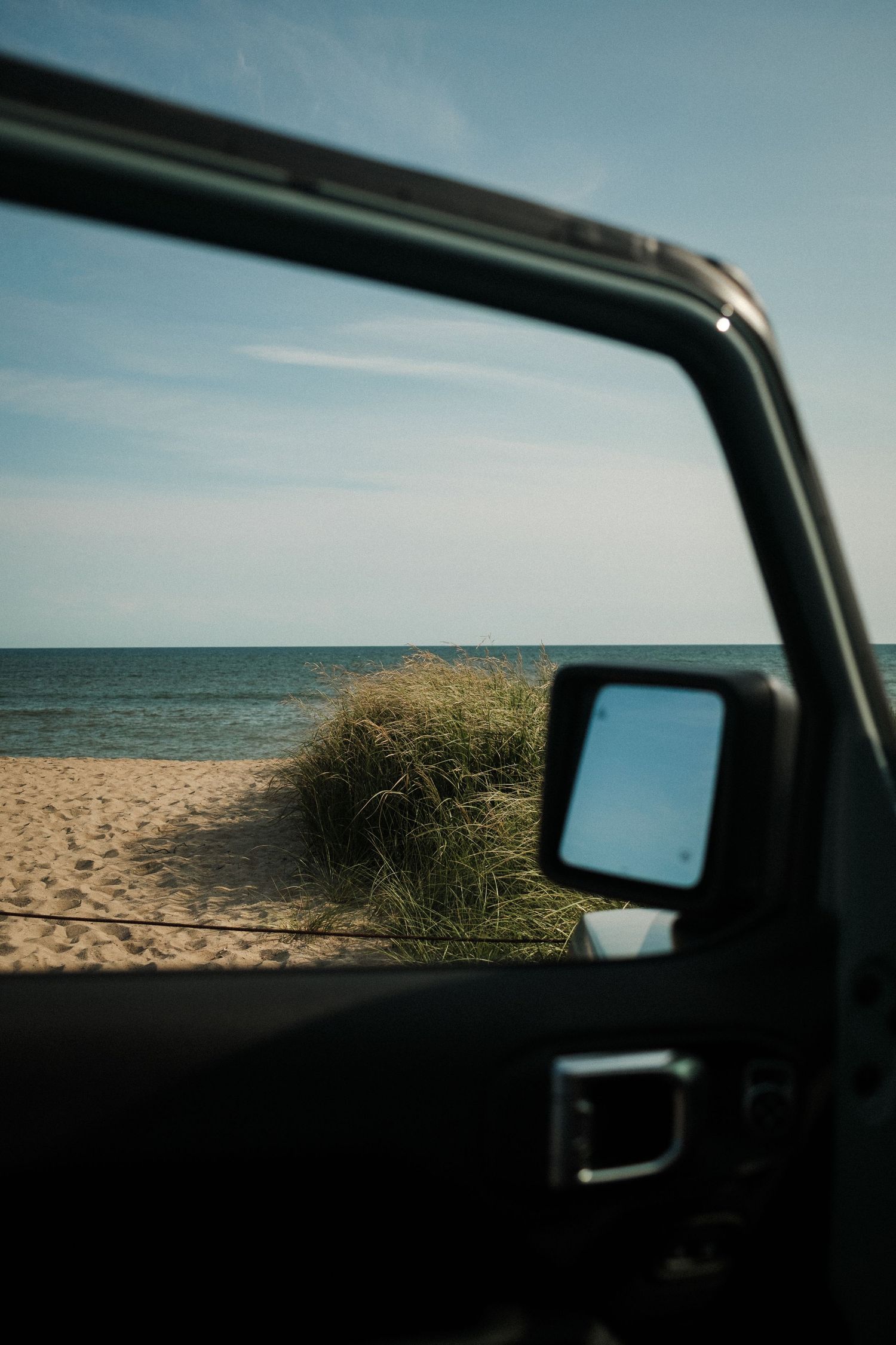 View of a sandy beach through a car window showing dunes and ocean in the distance.
