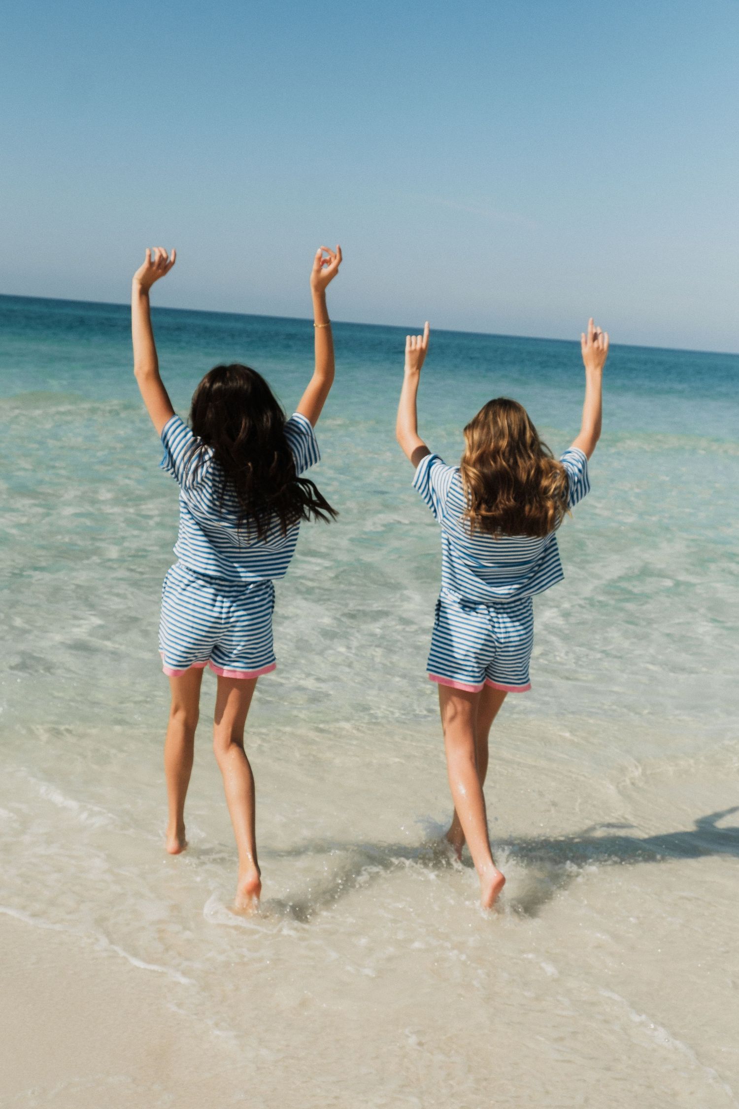 Two people in striped outfits jumping joyfully in shallow ocean water at the beach.
