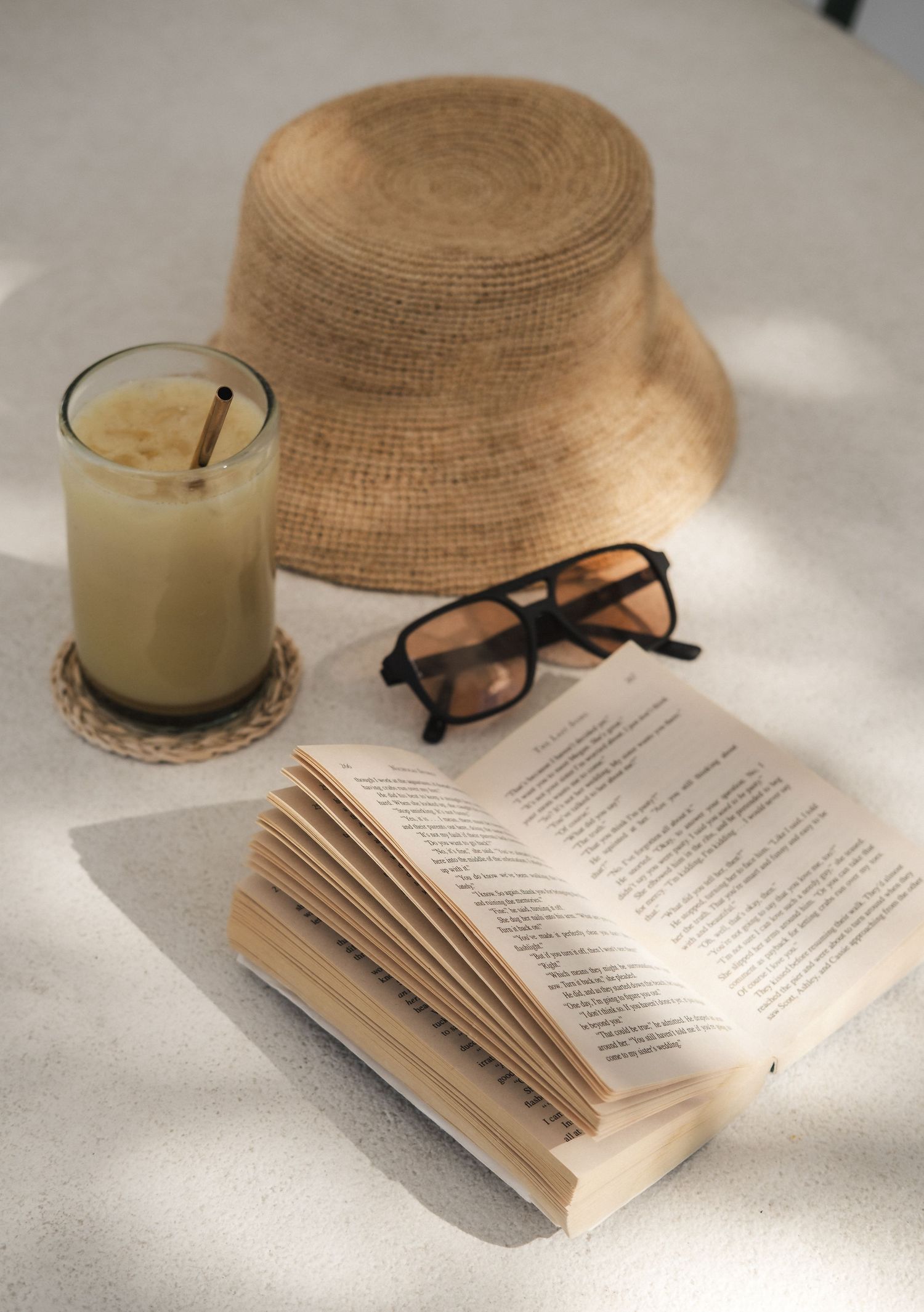 Cozy reading setup with straw hat, sunglasses, book and creamy drink in natural lighting.