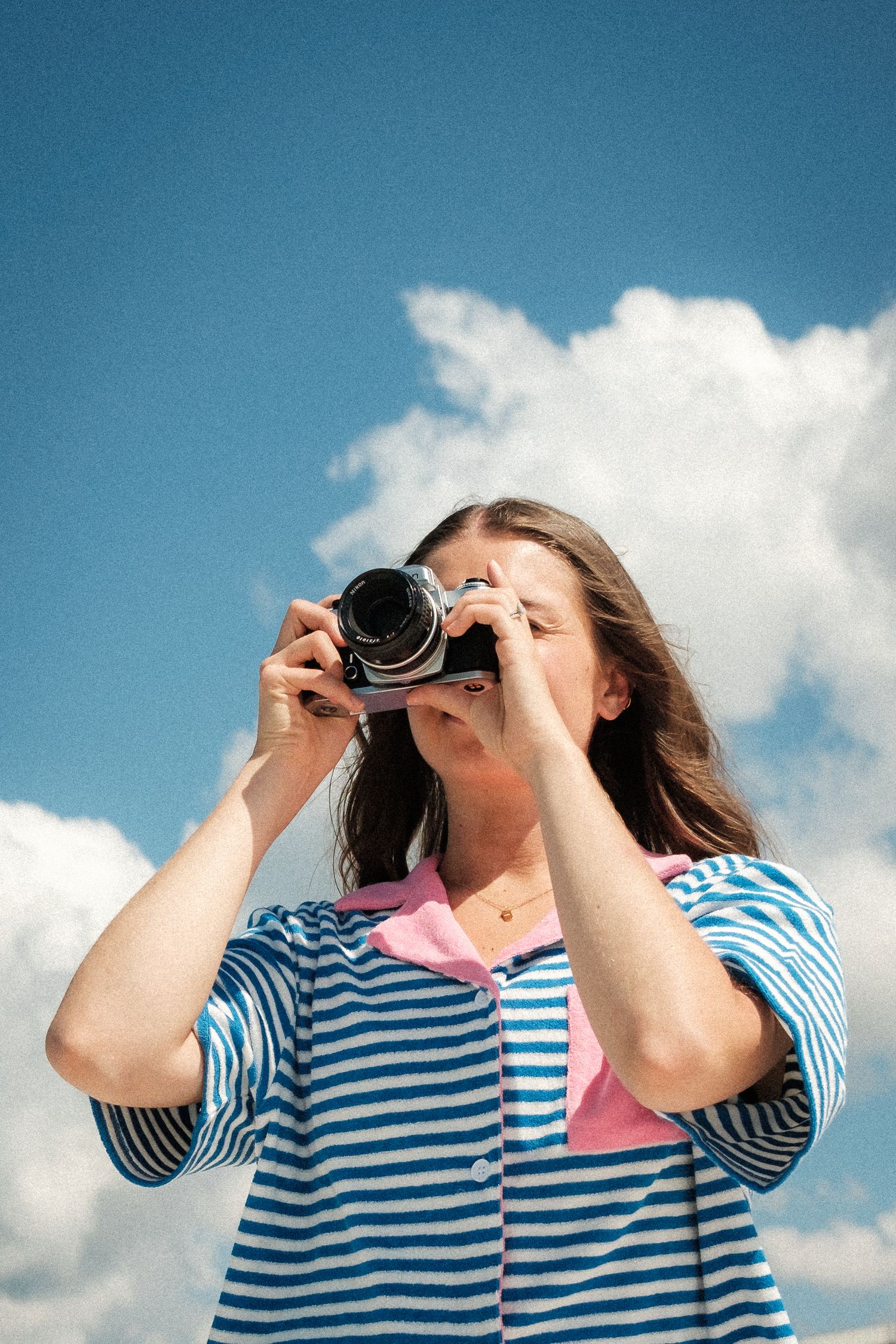 Person in striped shirt using binoculars against blue sky with white clouds.
