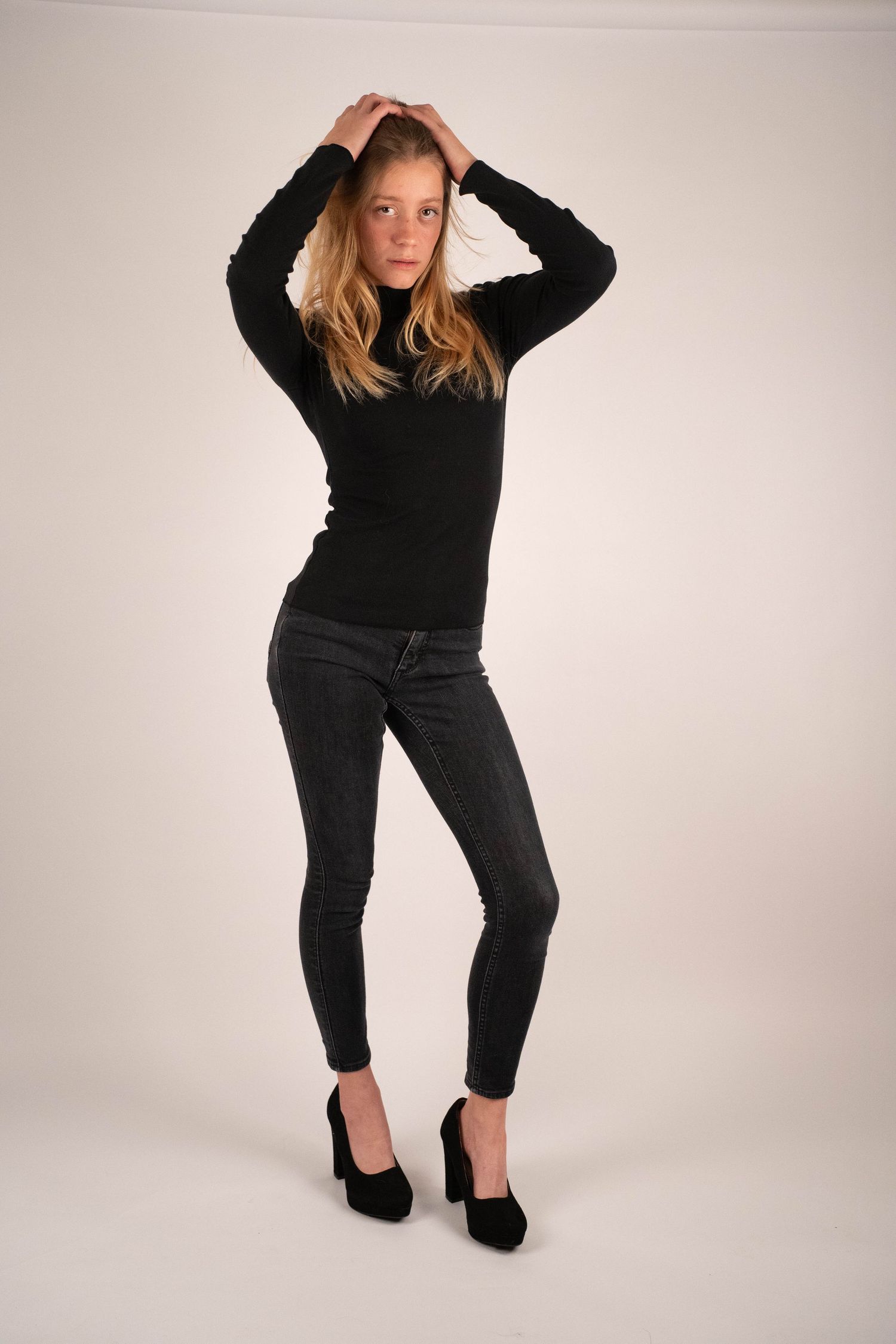 Blonde woman in black turtleneck and gray skinny jeans posing with hands in hair on white backdrop.