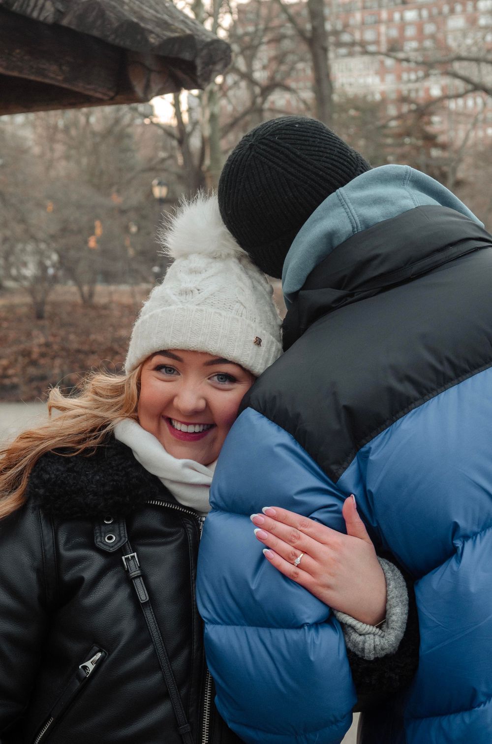 A couple in winter attire embracing outdoors on a cold day with one wearing a white knit hat and the other in a blue puffer coat.