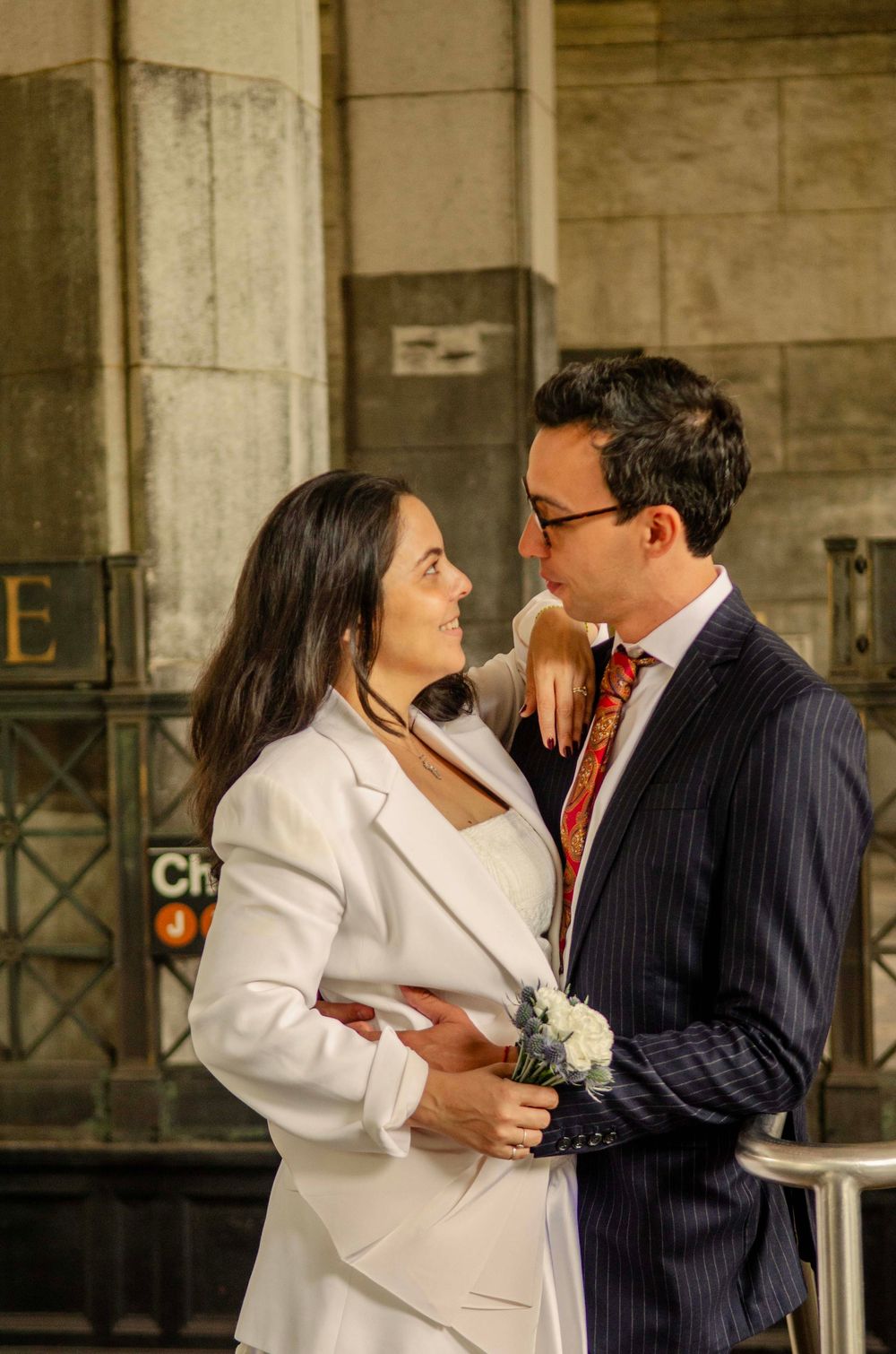 Couple in formal attire sharing a romantic moment near a NYC subway entrance, woman in white blazer holding flowers.