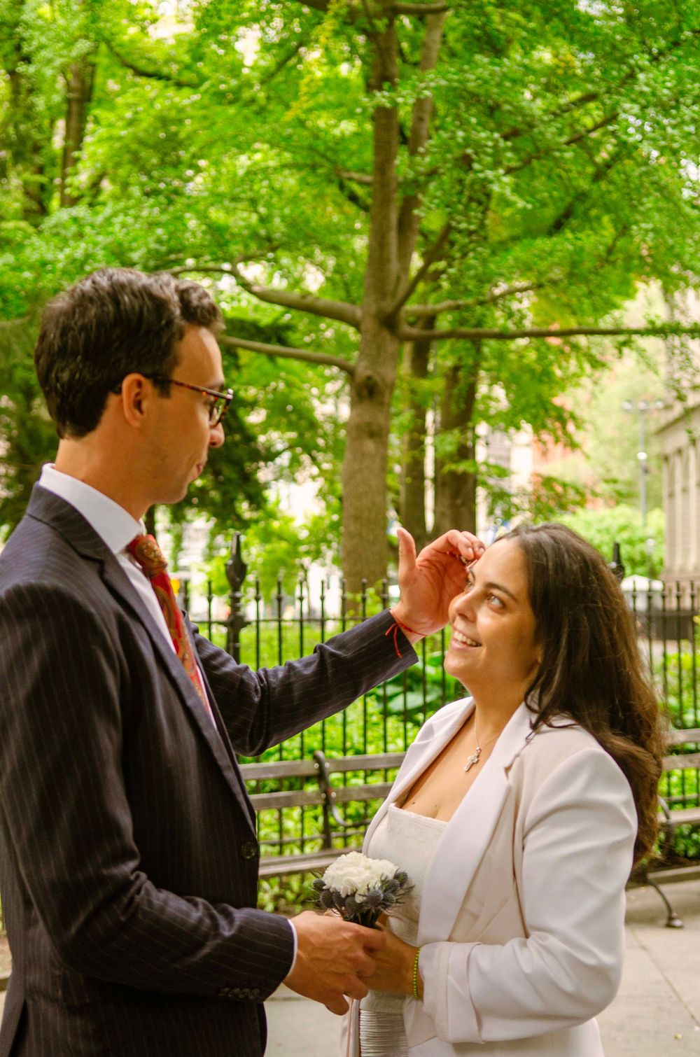 A groom in a dark suit gently touches his bride's face outdoors near a lush green park during their wedding day.