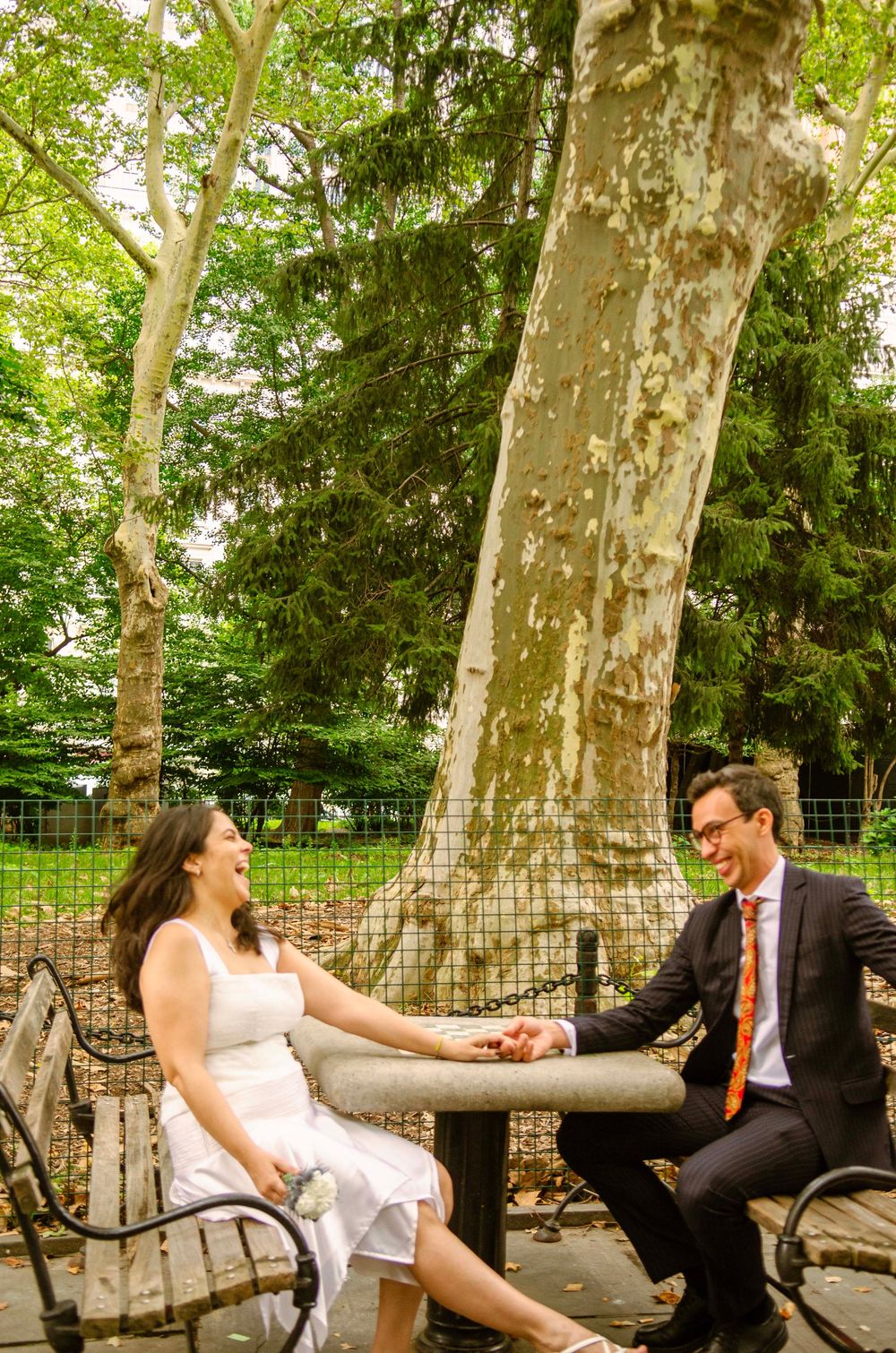 A smiling bride and groom hold hands on a park bench surrounded by lush trees on their wedding day.