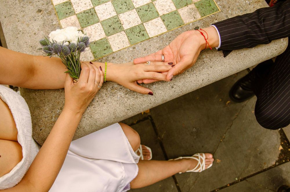 Bride in white dress holds flowers while groom in suit places ring on her finger on checkered steps.