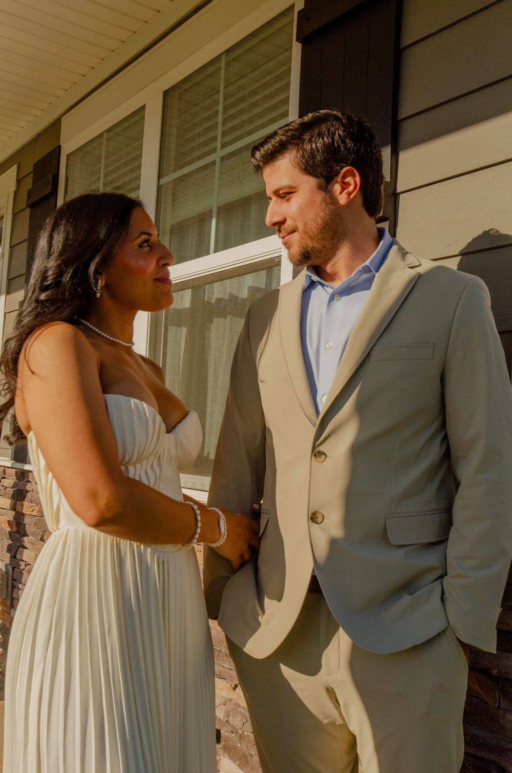 A couple in wedding attire share an intimate moment on a sunlit porch, with the bride in white and groom in gray suit.