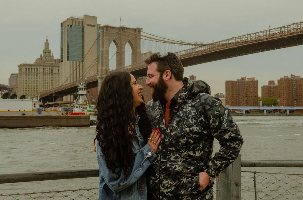 Couple sharing a romantic moment by the Brooklyn Bridge waterfront in New York City on a cloudy day.