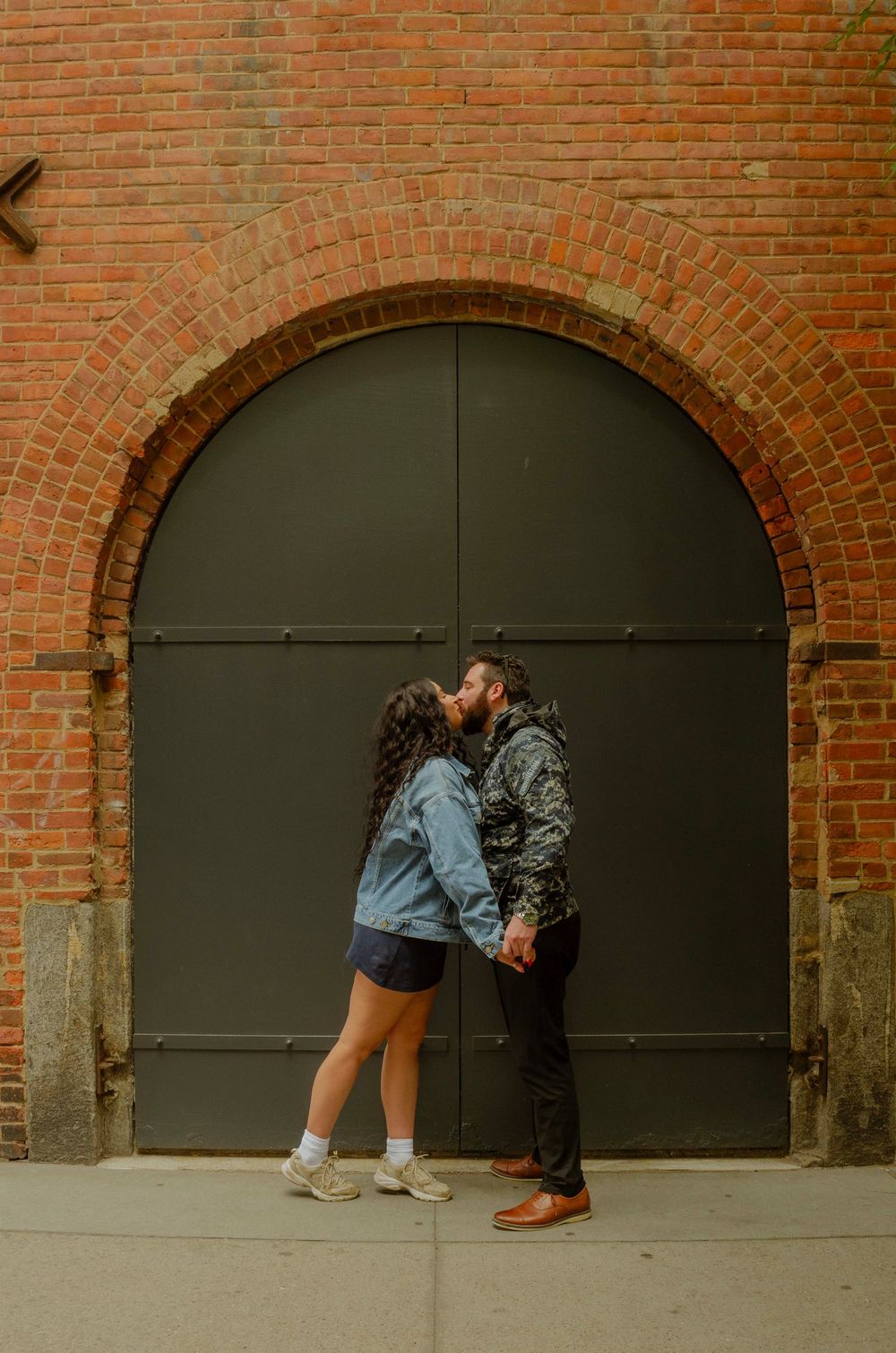 Couple sharing a romantic kiss in front of a large arched green door set in a red brick wall.
