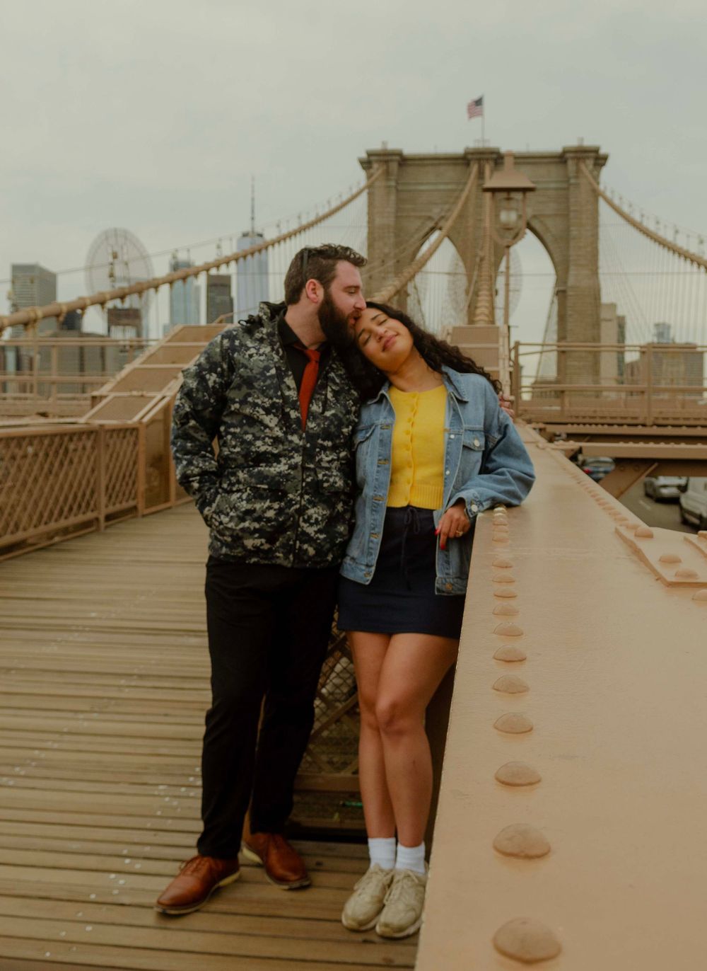 Couple sharing a romantic moment on the Brooklyn Bridge walkway with NYC skyline in the background.
