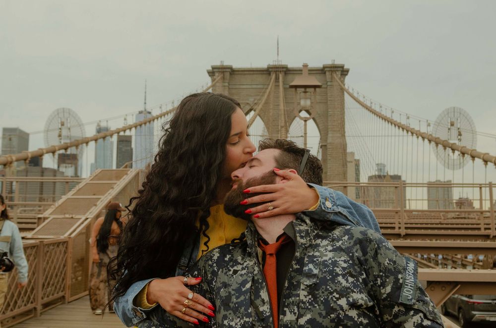 Couple sharing a kiss on the Brooklyn Bridge with NYC skyline in the background on a cloudy day.