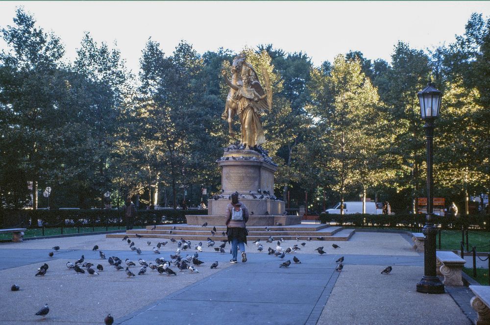 A stone monument statue stands in a park plaza surrounded by pigeons on a sunny day.