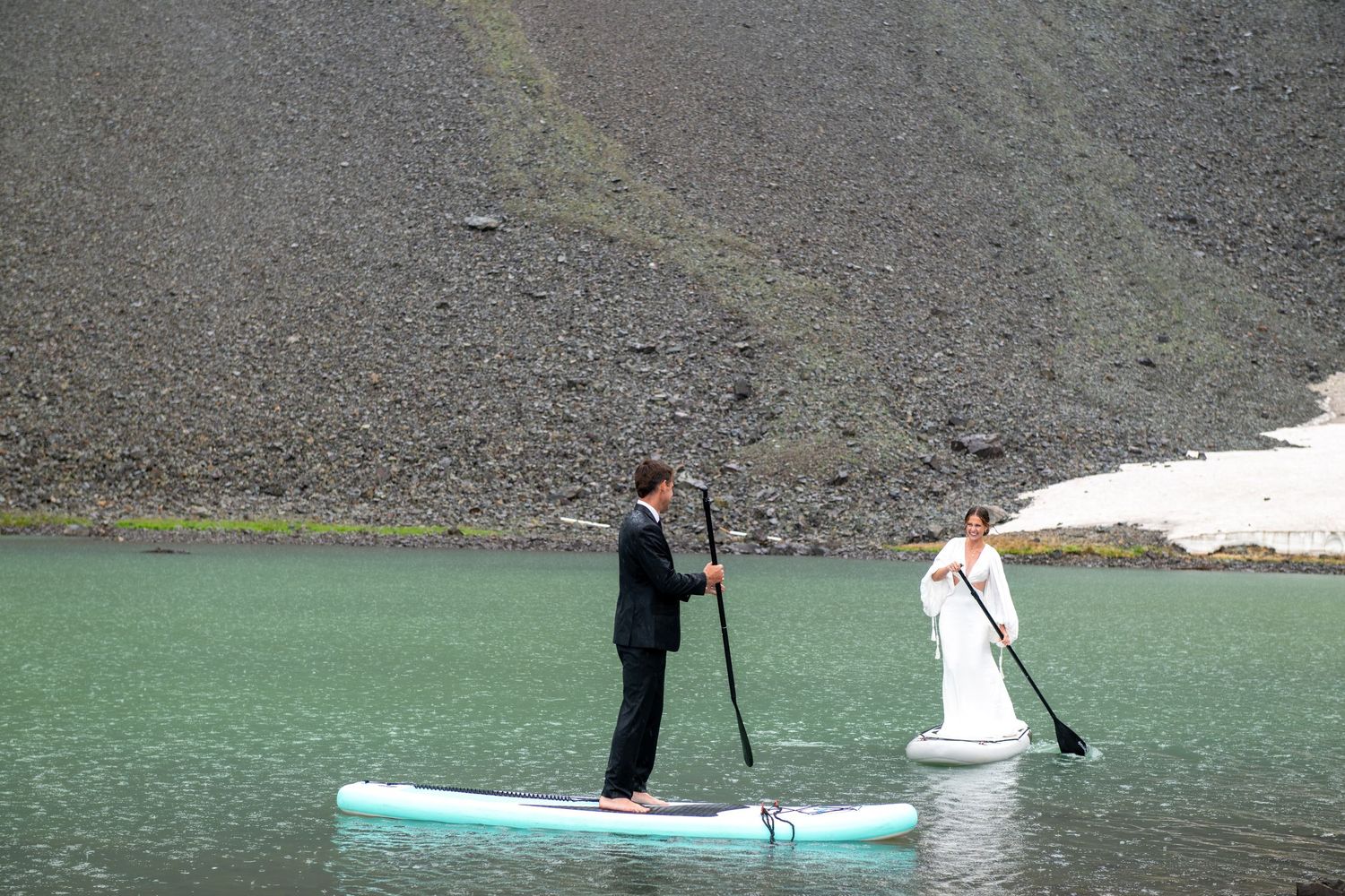 Bride and groom share a romantic moment on rocky shore with mountain lake and dramatic peaks backdrop.