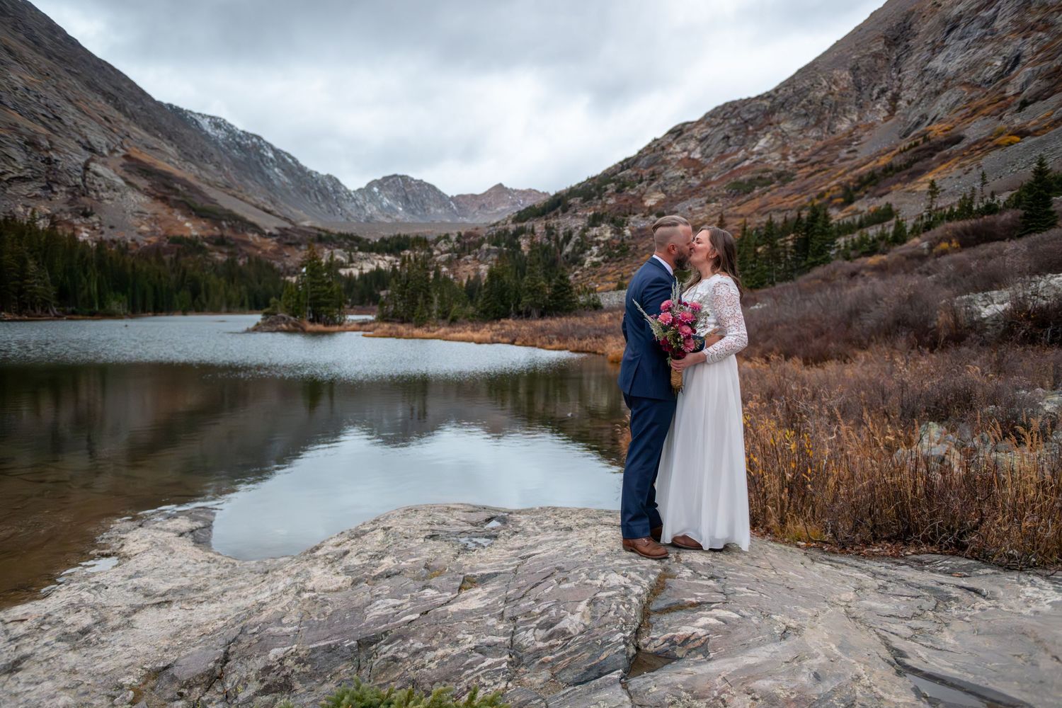 Bride and groom share a romantic moment on rocky shore with mountain lake and dramatic peaks backdrop.