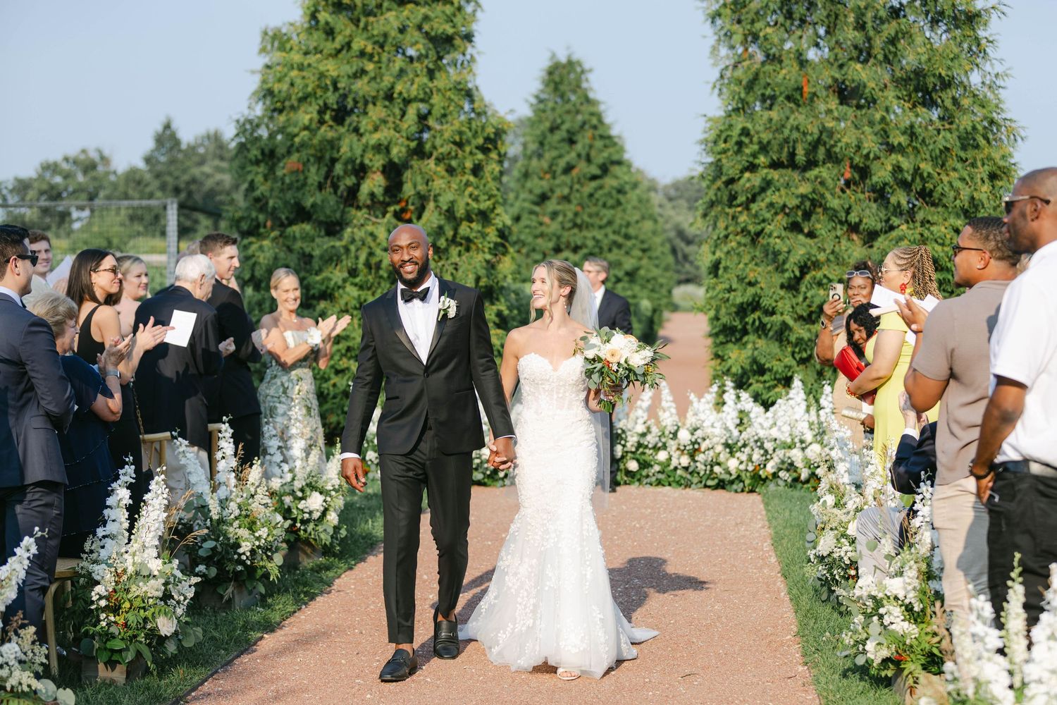 A newlywed couple walks down an outdoor aisle lined with white flowers as guests celebrate at a garden ceremony. - Elawa Farm
