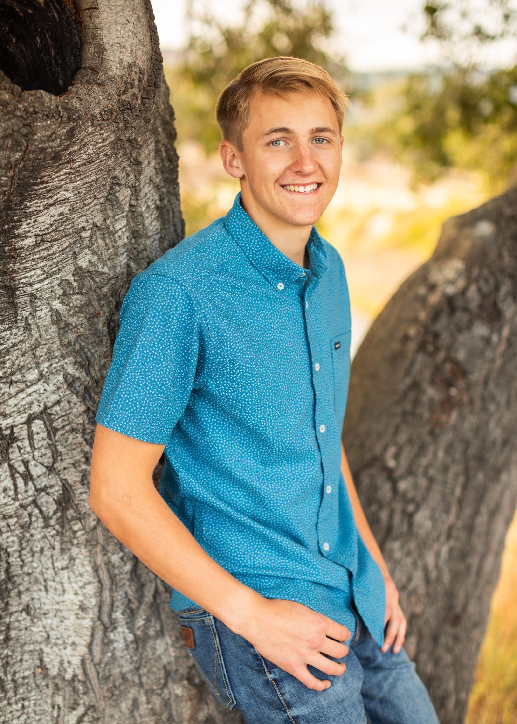 Person wearing bright turquoise button-down shirt poses against large tree trunk in outdoor portrait session.