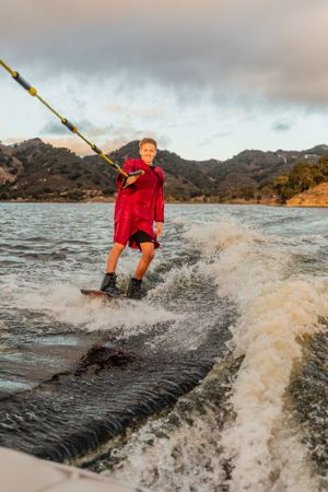 Person wakeboarding in red outfit creates splash on lake with mountains in background.