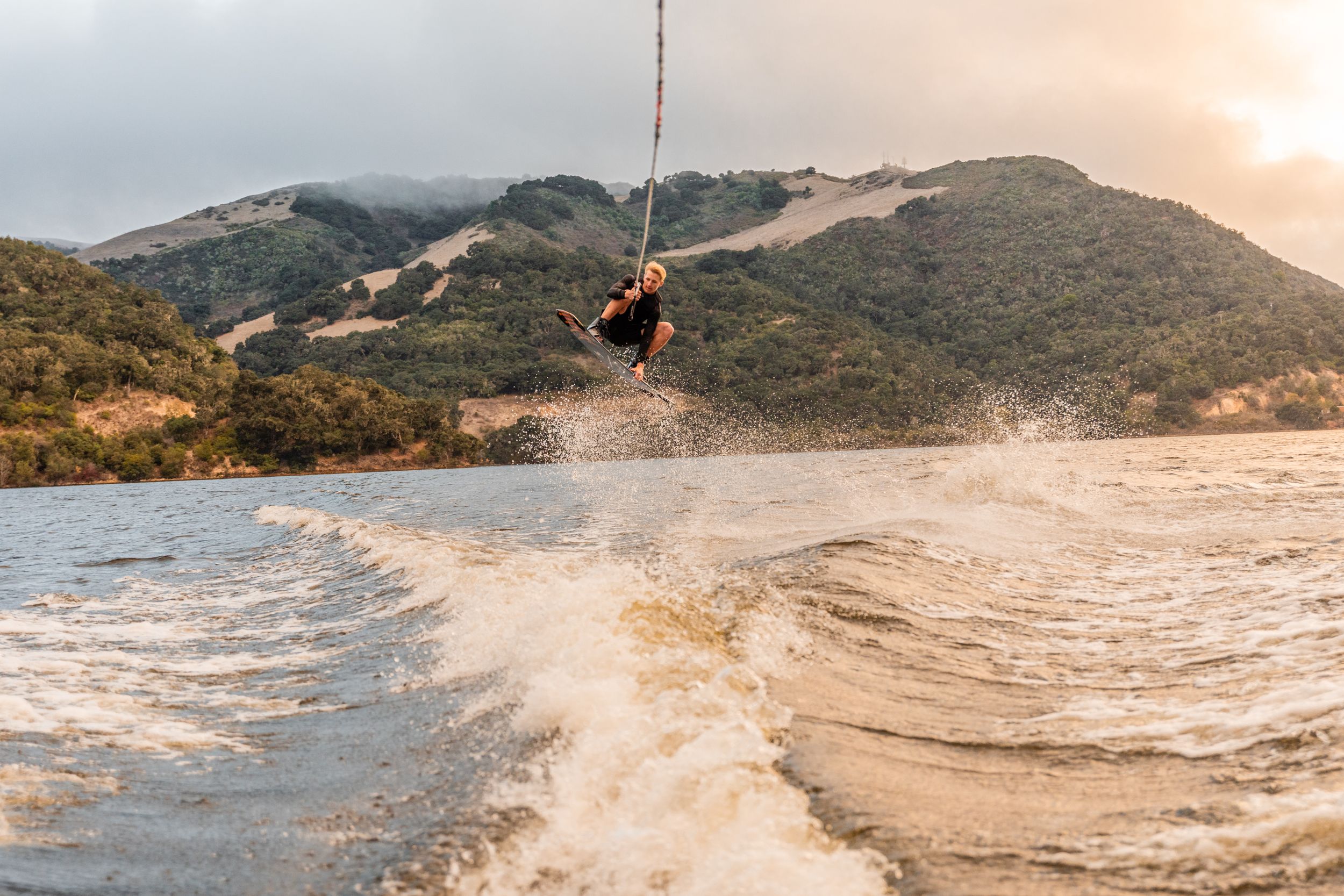 Action shot of wakeboarding jump silhouetted against mountain landscape at sunset.
