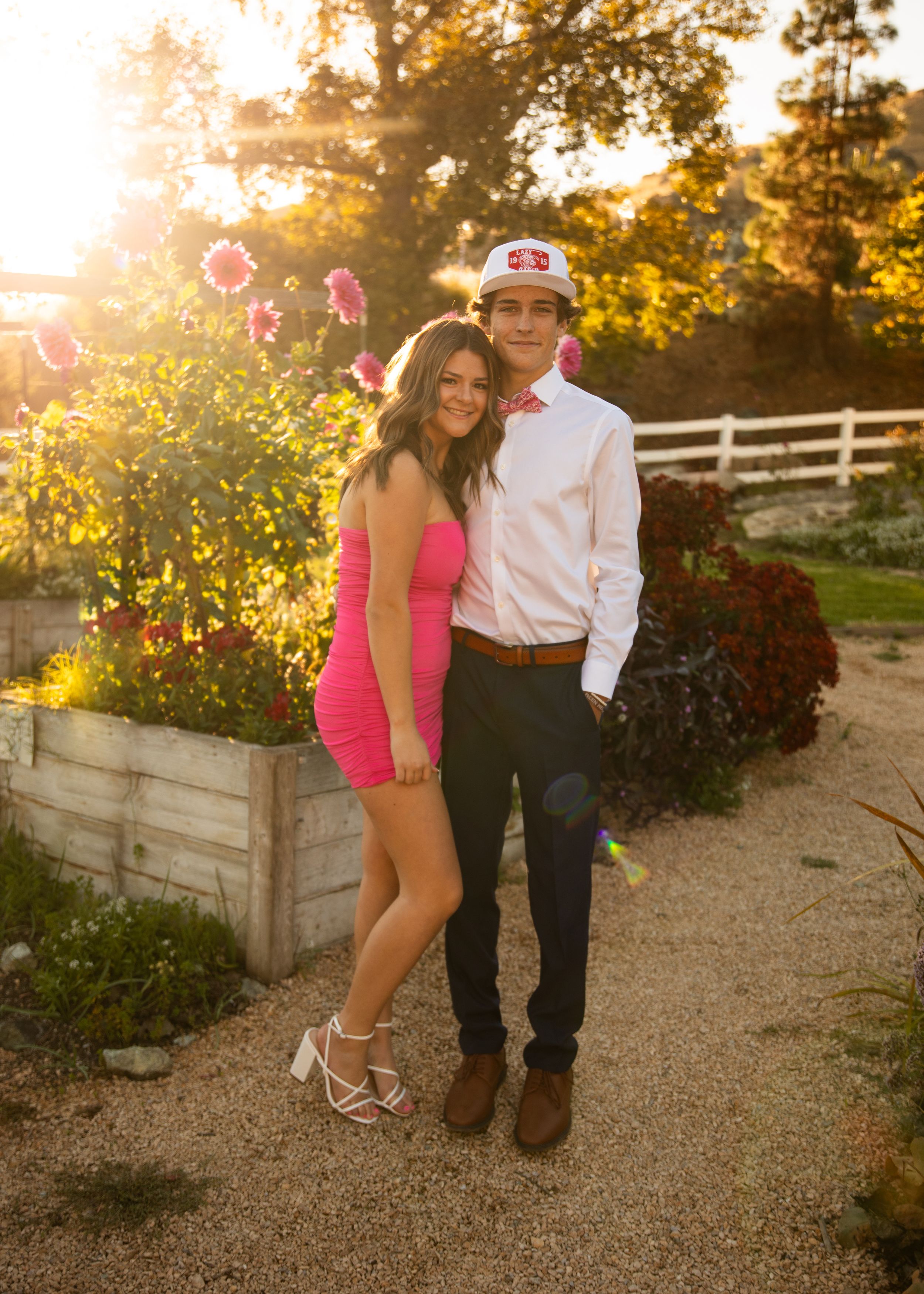 Couple poses together in garden at sunset with pink flowers and white fence in background.