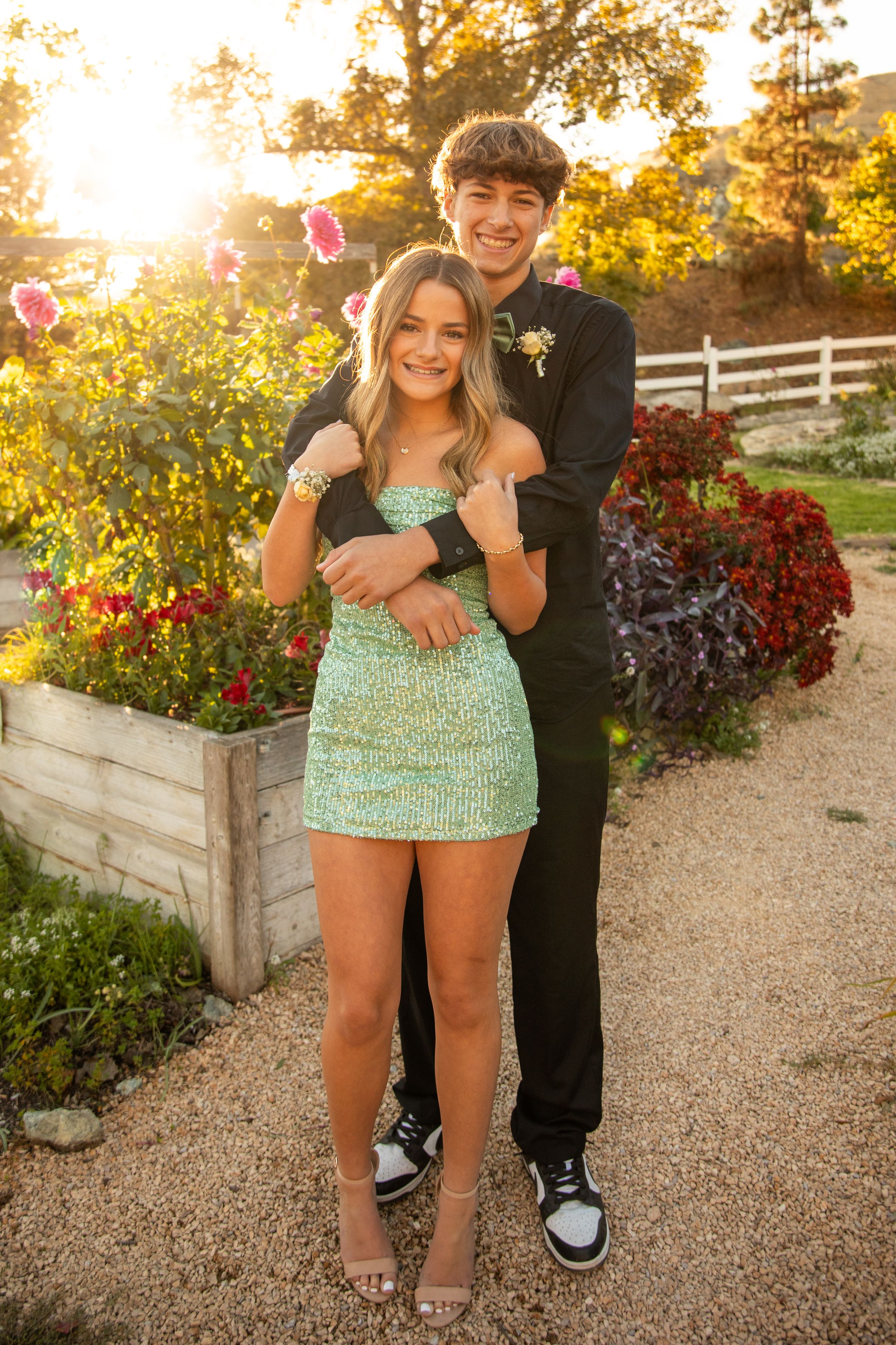 Elegant formal couple embraces near flowering garden during golden hour.