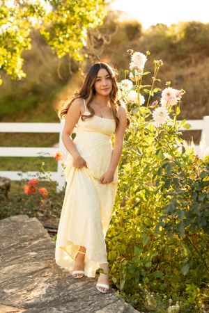 A woman in pale yellow dress poses by blooming garden flowers in warm evening light.