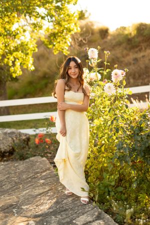 Woman in yellow dress poses by blooming flowers during golden hour sunset in garden setting.