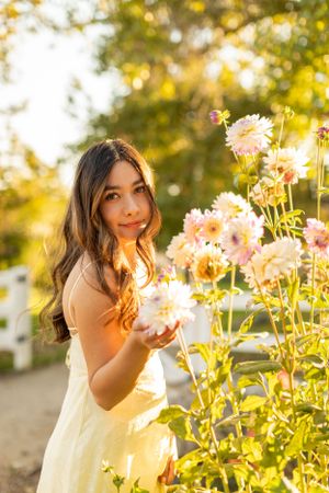 Woman in pale dress admires flowers in sunny garden with warm evening light.
