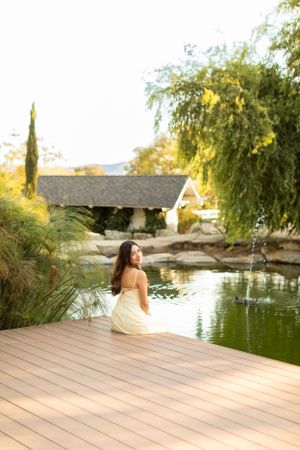 Figure in dress sits on wooden deck by serene pond at sunset with willow trees.