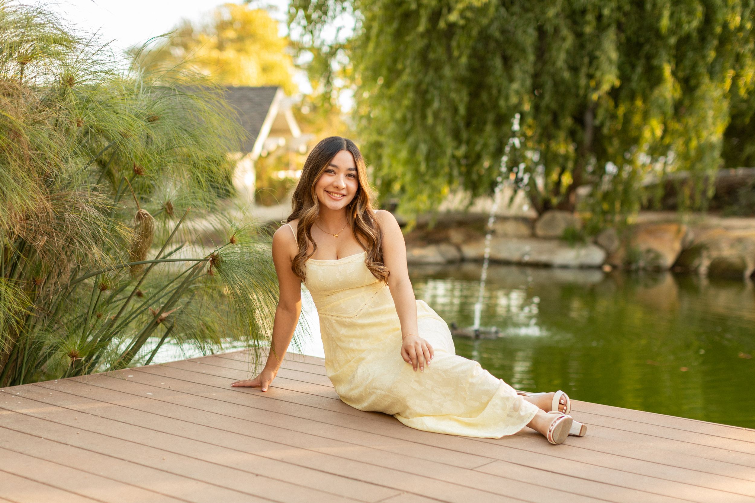 Woman in pale dress reclines on wooden deck by tranquil pond at golden hour.