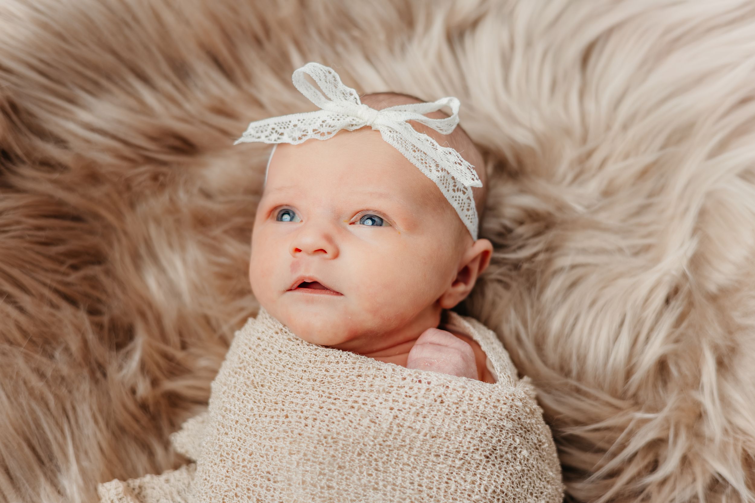 Baby wearing white lace headband lies on soft beige fur blanket wrapped in neutral swaddle.