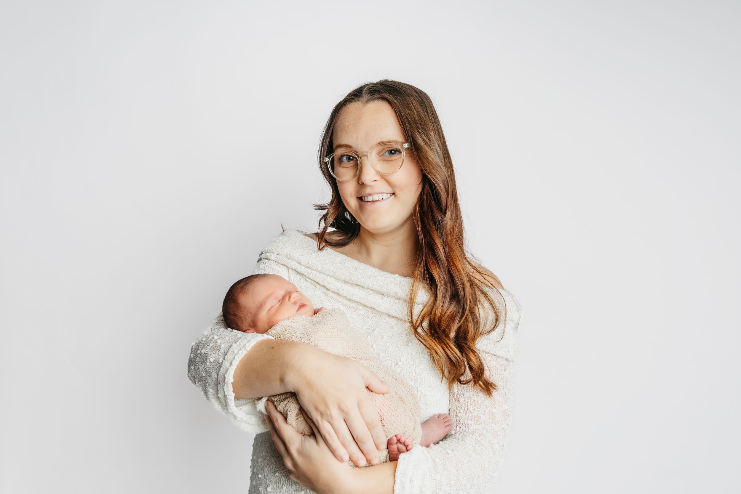 Woman in white sweater holds sleeping newborn against white background.