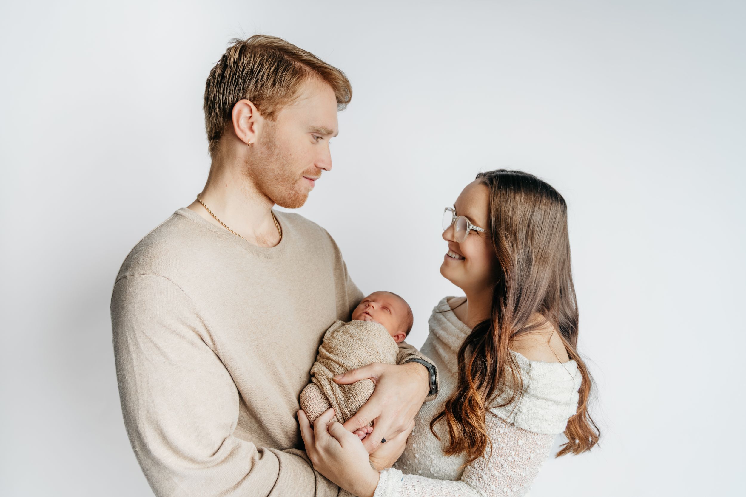 Parents in neutral colored clothing tenderly holding their newborn baby against white background.