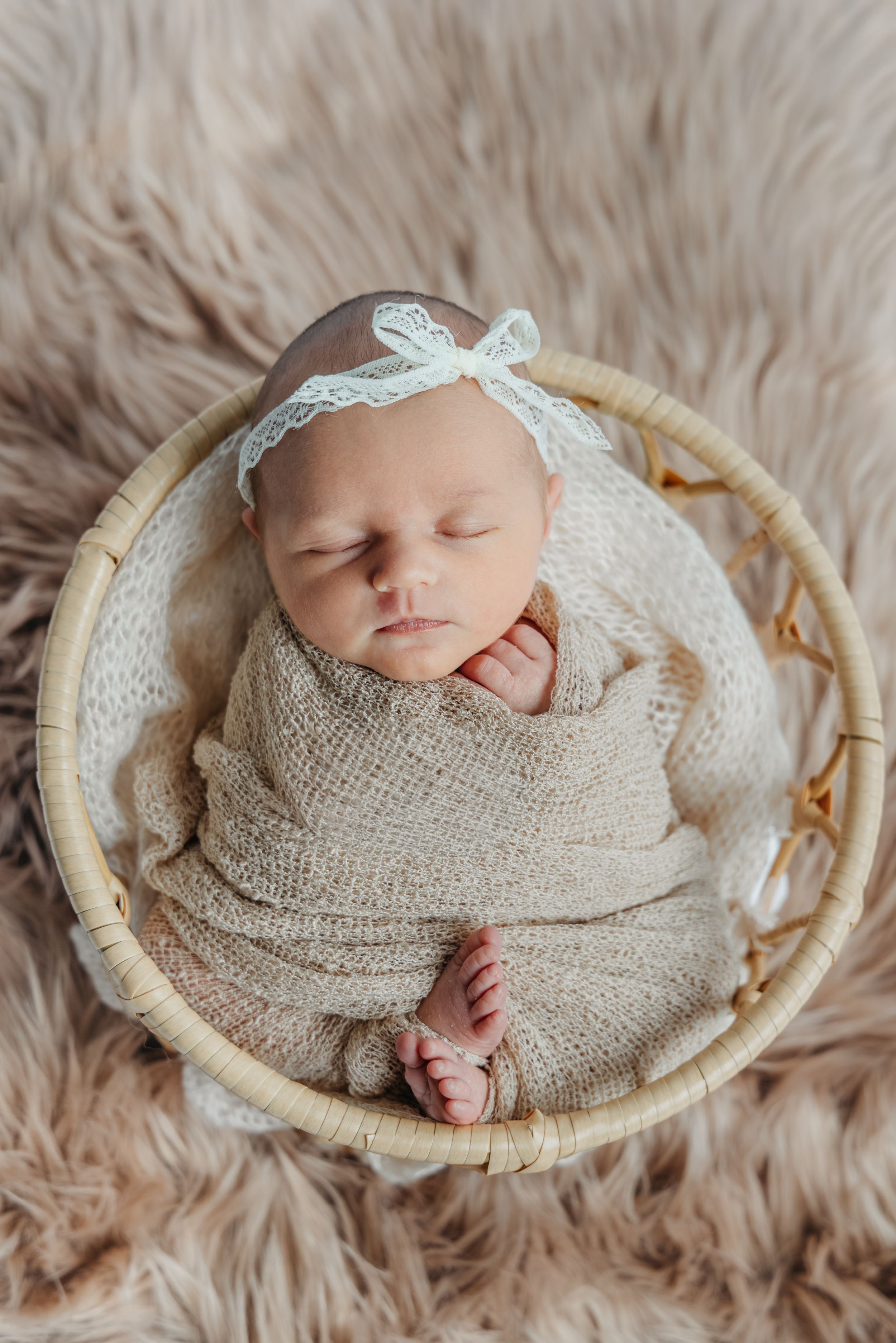 Sleeping newborn baby wrapped in beige knit blanket on round basket with furry backdrop.
