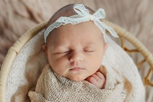 Sleeping newborn baby wrapped in white lace swaddle with matching lace bonnet resting on cream blanket.