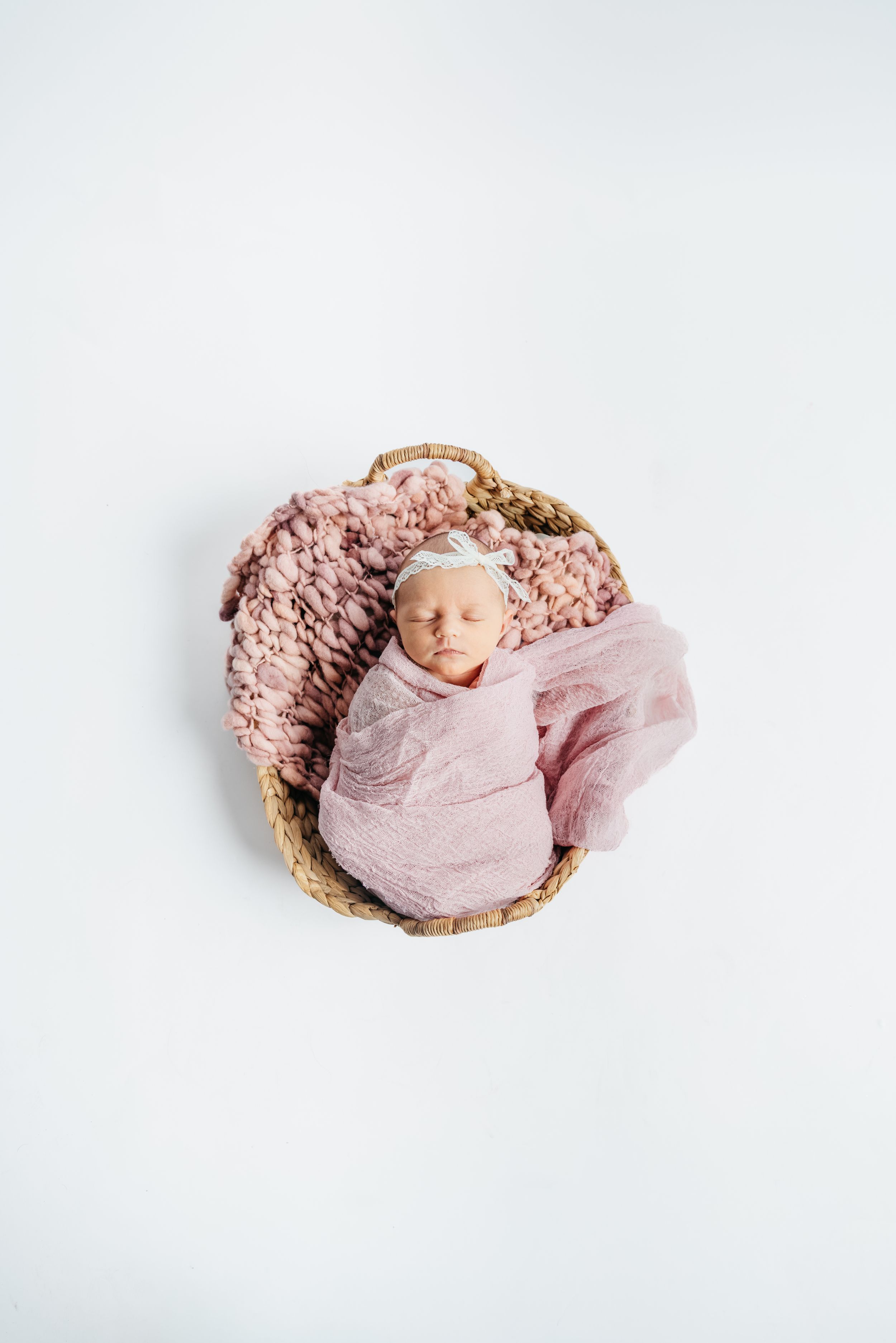 A sleeping newborn wrapped in soft pink fabric rests peacefully in a round woven basket on white background.