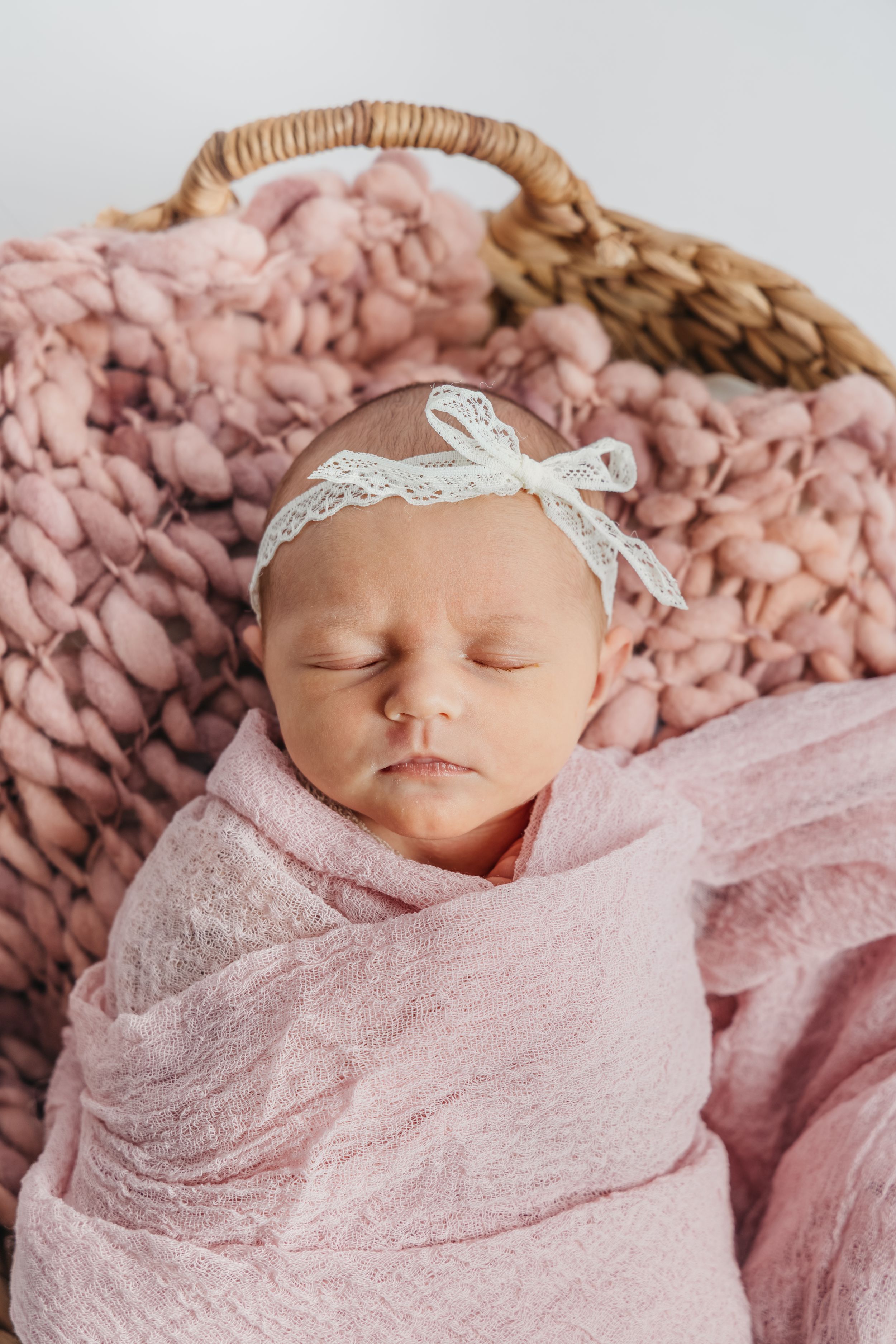 Sleeping newborn baby wrapped in soft pink lace swaddle with delicate white headband on textured pink blanket.