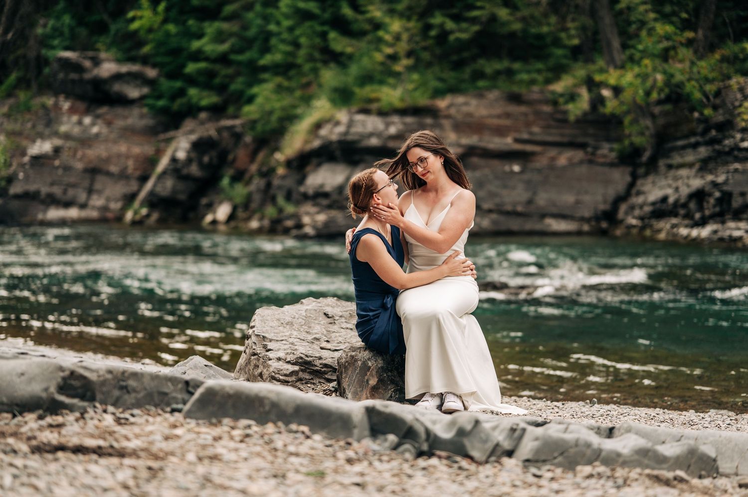 A couple embraces on rocky riverbank surrounded by lush forest scenery during a romantic outdoor photoshoot.