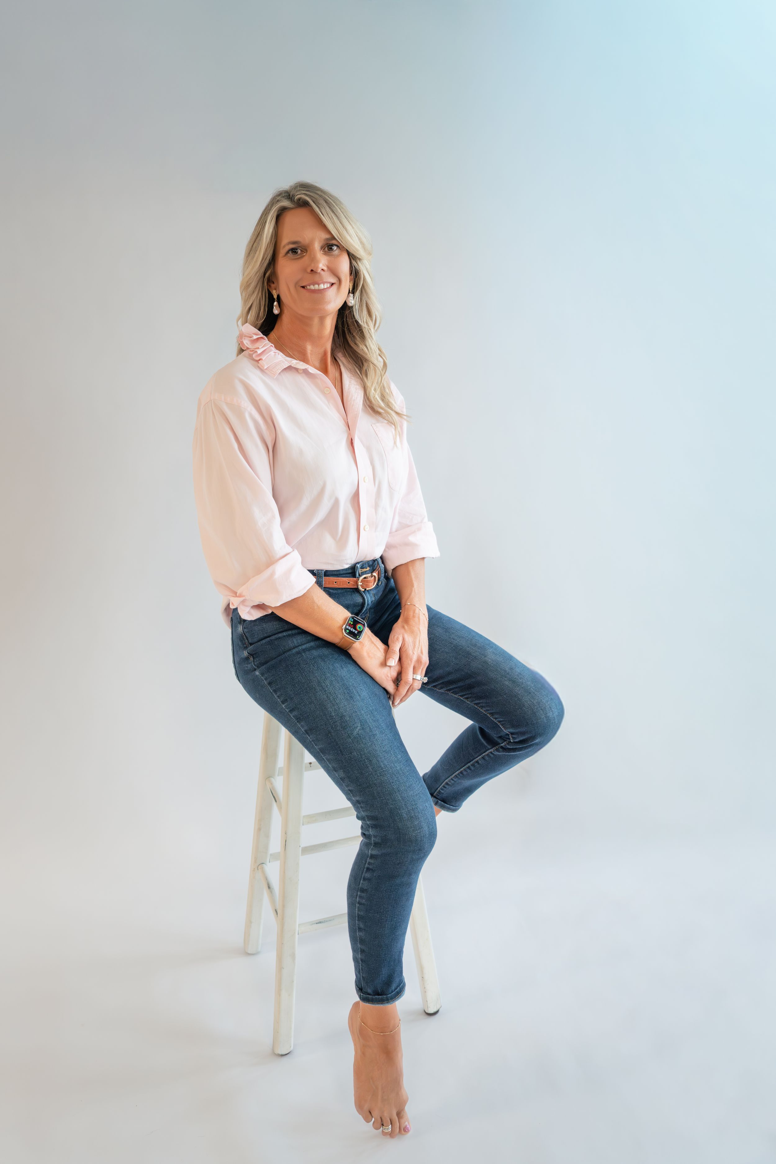 Smiling blonde woman in pink shirt and blue jeans sitting on a white stool against a light gray background.