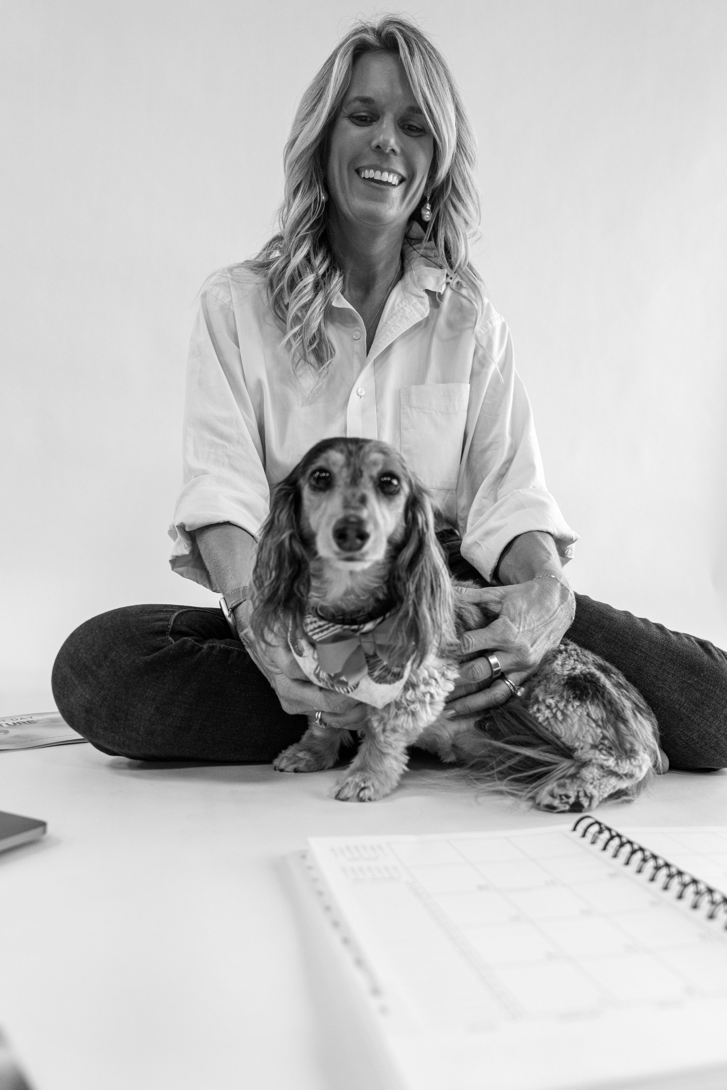 Smiling woman in white shirt sitting cross-legged with a dachshund dog, black and white photo with notebook in foreground.