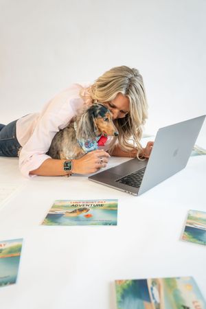 Blonde woman lying on white floor with dachshund dog, working on MacBook laptop surrounded by adventure-themed books.
