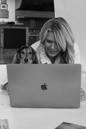 Black and white photo of a blonde woman smiling at a MacBook laptop with a dachshund dog beside her.