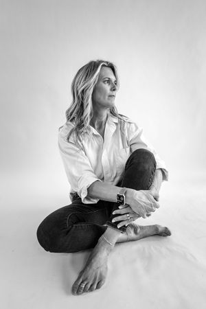 Black and white portrait of a blonde woman sitting cross-legged on floor in white shirt and jeans, gazing sideways.