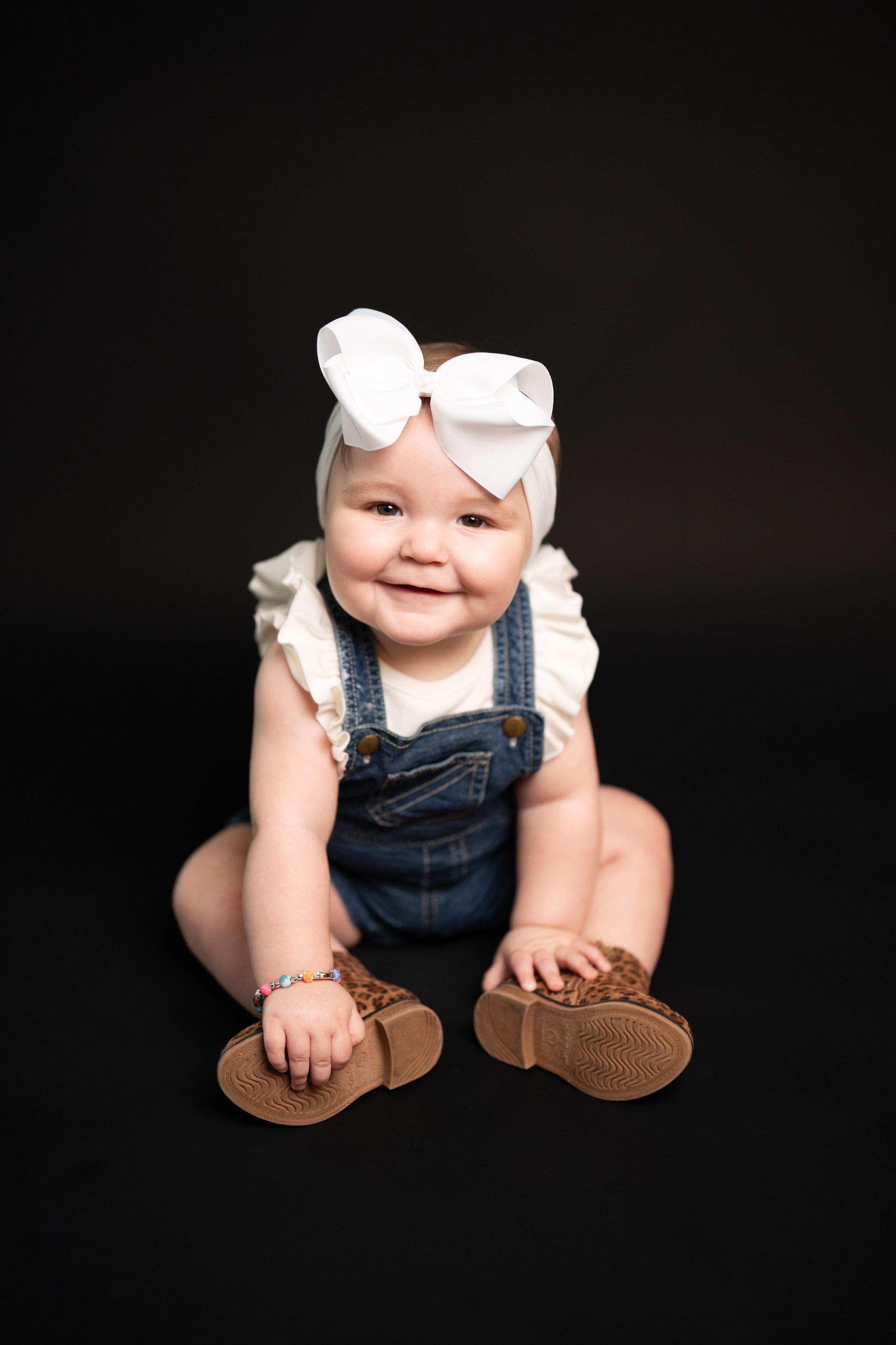 Baby wearing denim overalls and white bow sits on black background showing adorable smile.
